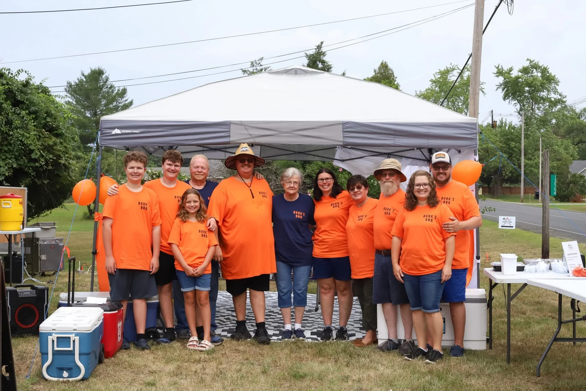Group of people, a family, standing under a tent at an outdoor BBQ event, wearing matching orange T-shirts with 'Burk's BBQ' printed on them.