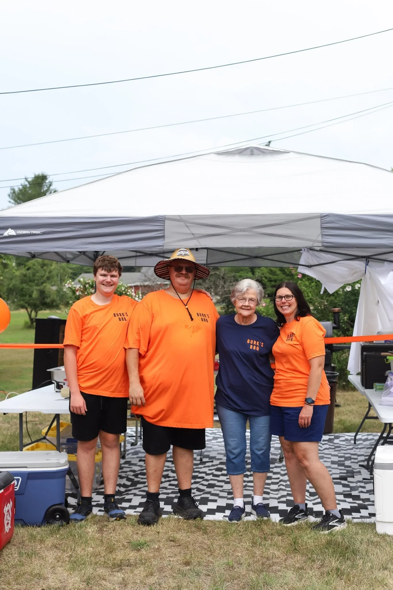 Group of people standing outdoors under a canopy tent. wearing bright orange shirts with Burk's BBQ 