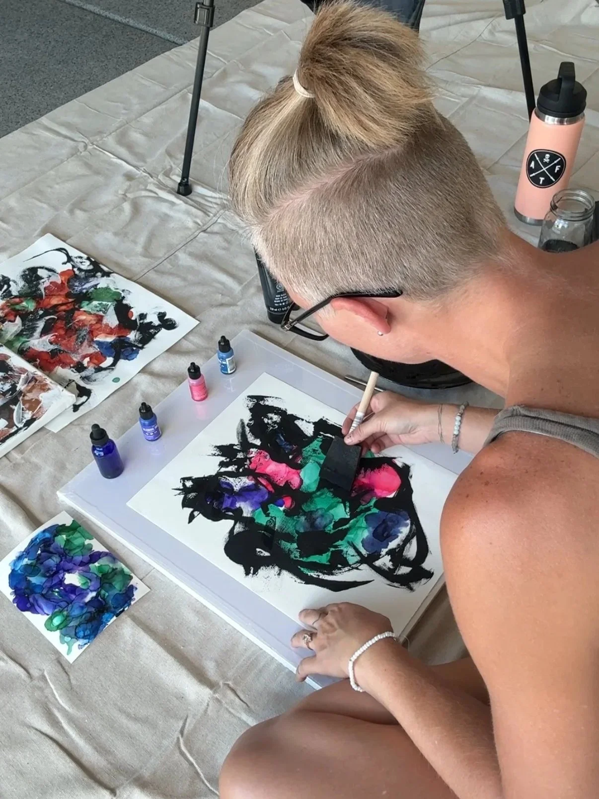 A woman with blonde hair tied in a bun and glasses creating an abstract watercolor painting on paper, surrounded by bottles of paint, a paintbrush, and a palette of colors, on a beige surface.