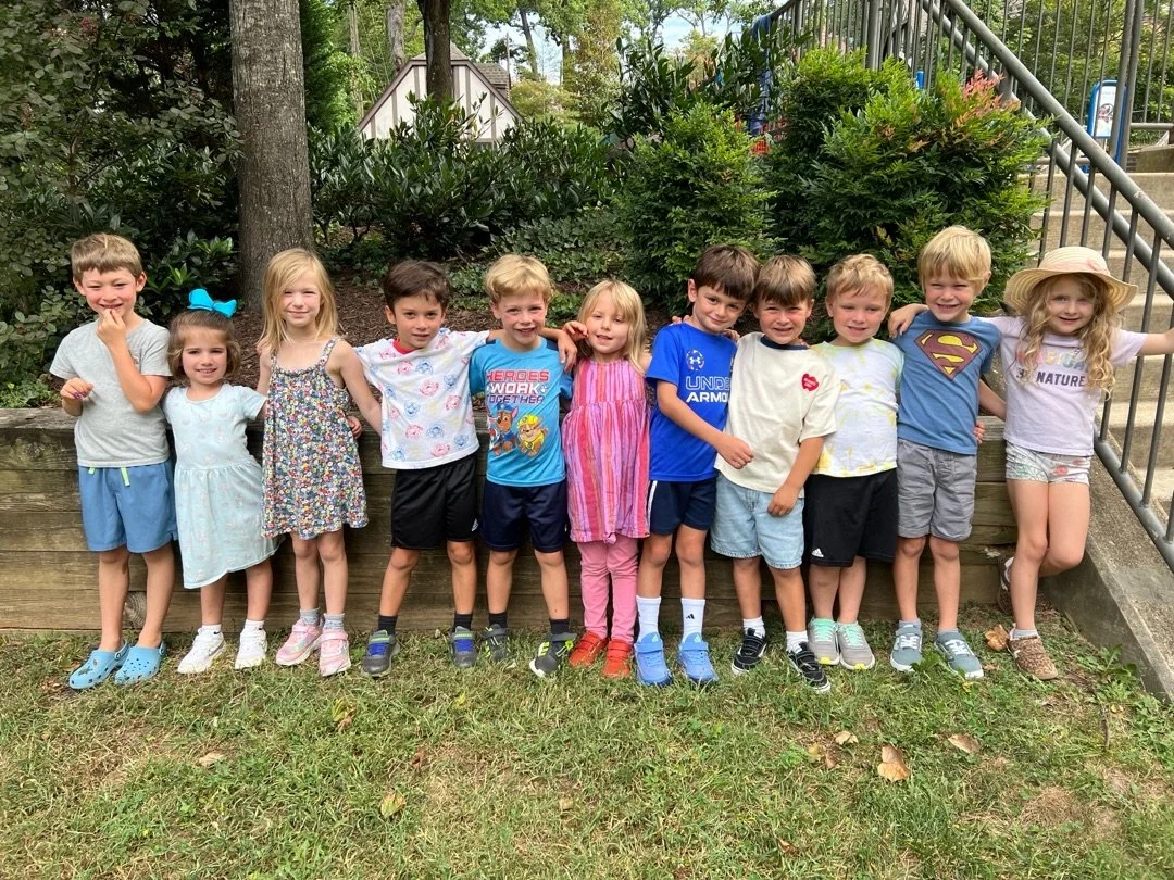 A group of fourteen children standing outdoors in front of bushes, smiling, with some holding each other.