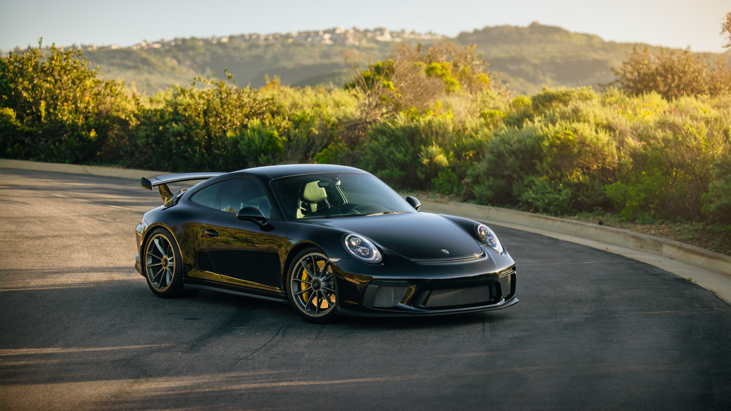 Black Porsche sports car parked on a paved road with green bushes and hills in the background during sunset.