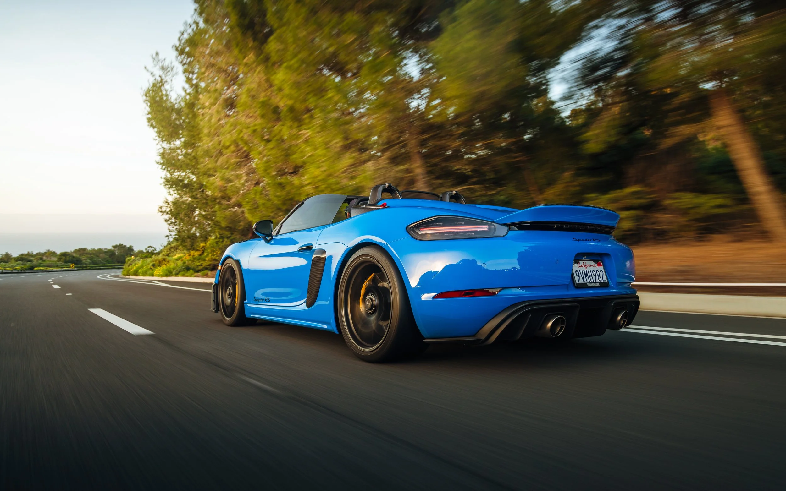Blue sports car driving on a rural open road with trees on the side and a view of the sky in the background.