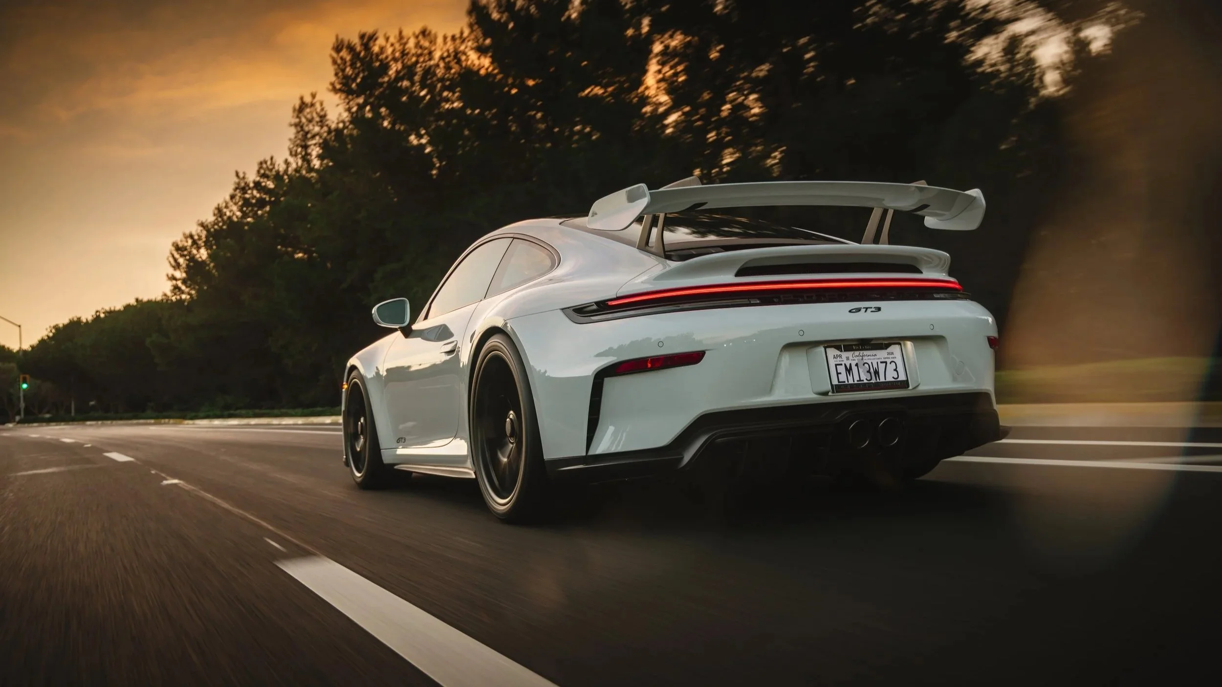 A white Porsche 911 GT3 driving on a highway at sunset, with a large rear wing and black wheels.
