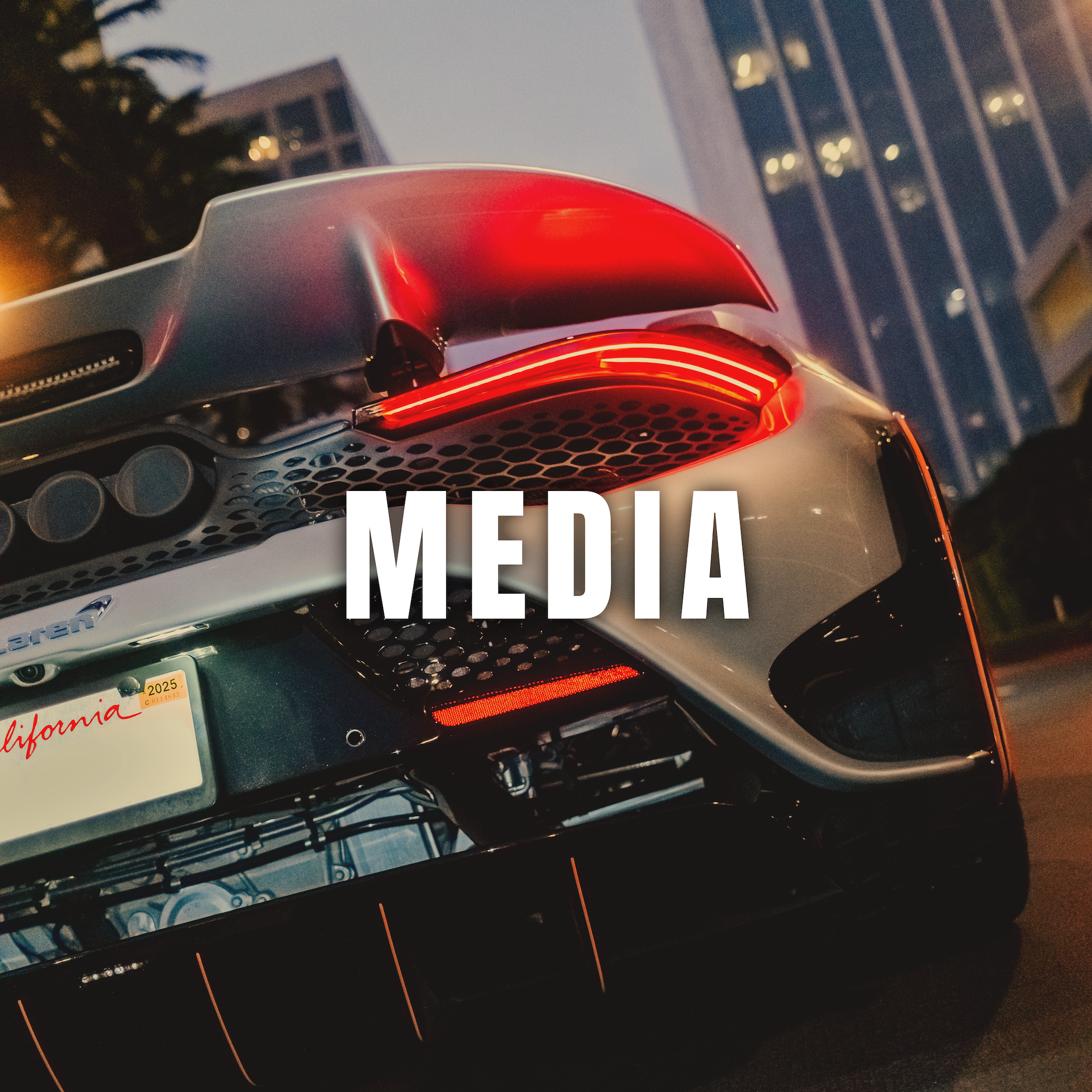 Close-up of the rear end of a high-performance sports car with illuminated tail lights and a custom license plate, city buildings in the background, and the word 'MEDIA' overlaid.