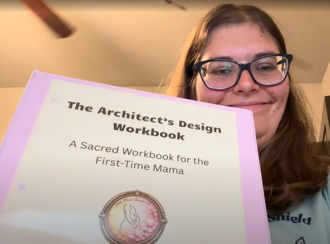 A woman with glasses and long brown hair holding a pink workbook titled "The Architect's Design Workbook: A Sacred Workbook for the First-Time Mama." She is smiling and taking a selfie in a room with a ceiling fan.