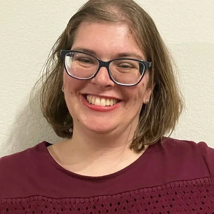 A woman with shoulder-length light brown hair, wearing glasses with black frames, smiling, and dressed in a maroon top with decorative stitching, standing against a plain light-colored wall.