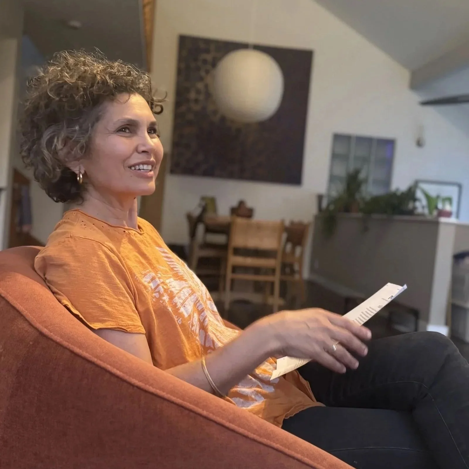 A woman with curly hair wearing an orange t-shirt, sitting on a sofa, holding a piece of paper, smiling in a cozy living room with wooden furniture and a large red and blue artwork on the wall.