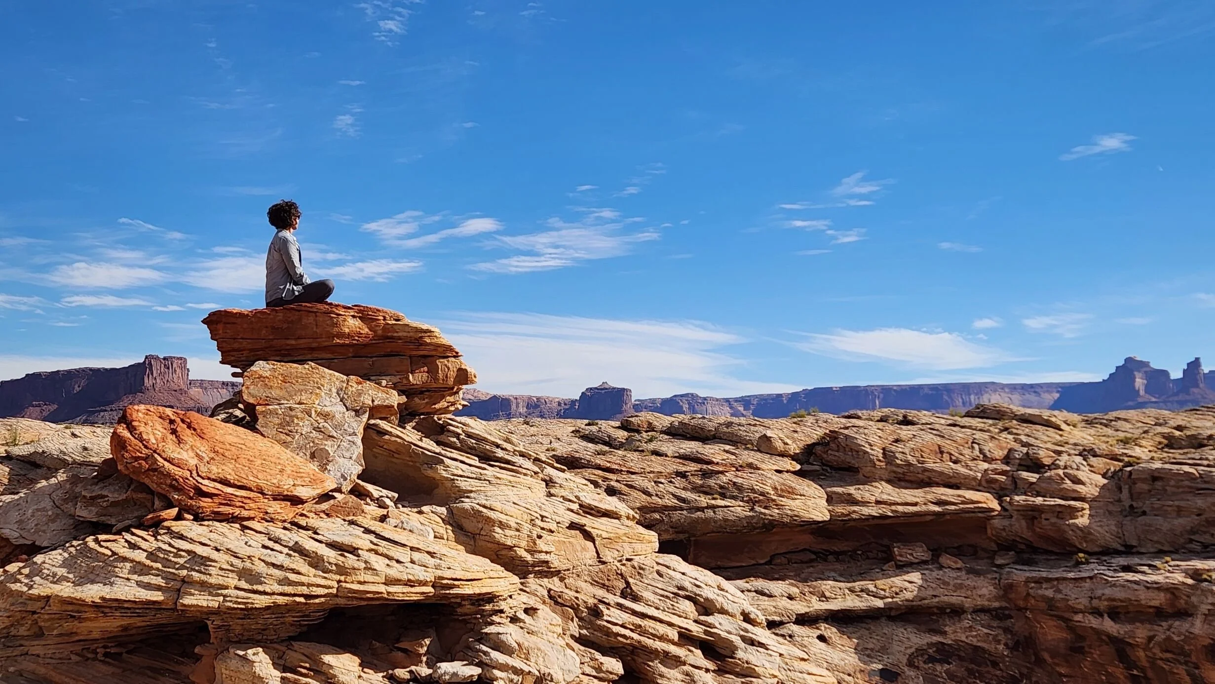 A person sitting cross-legged on a rock formation in a desert landscape, with blue sky and distant mesas in the background.