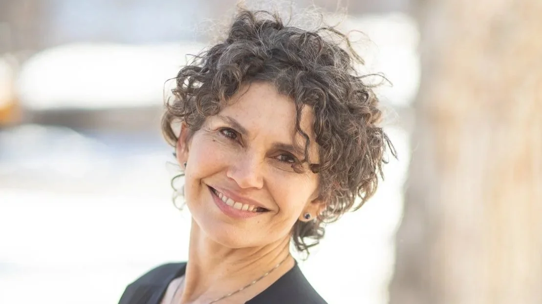 A smiling woman with short curly hair in an outdoor setting.