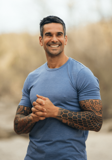 Stefanos Sifandos  with short dark hair and tattoos on his arms, wearing a blue t-shirt, standing outdoors with a blurred background of trees and an overcast sky.