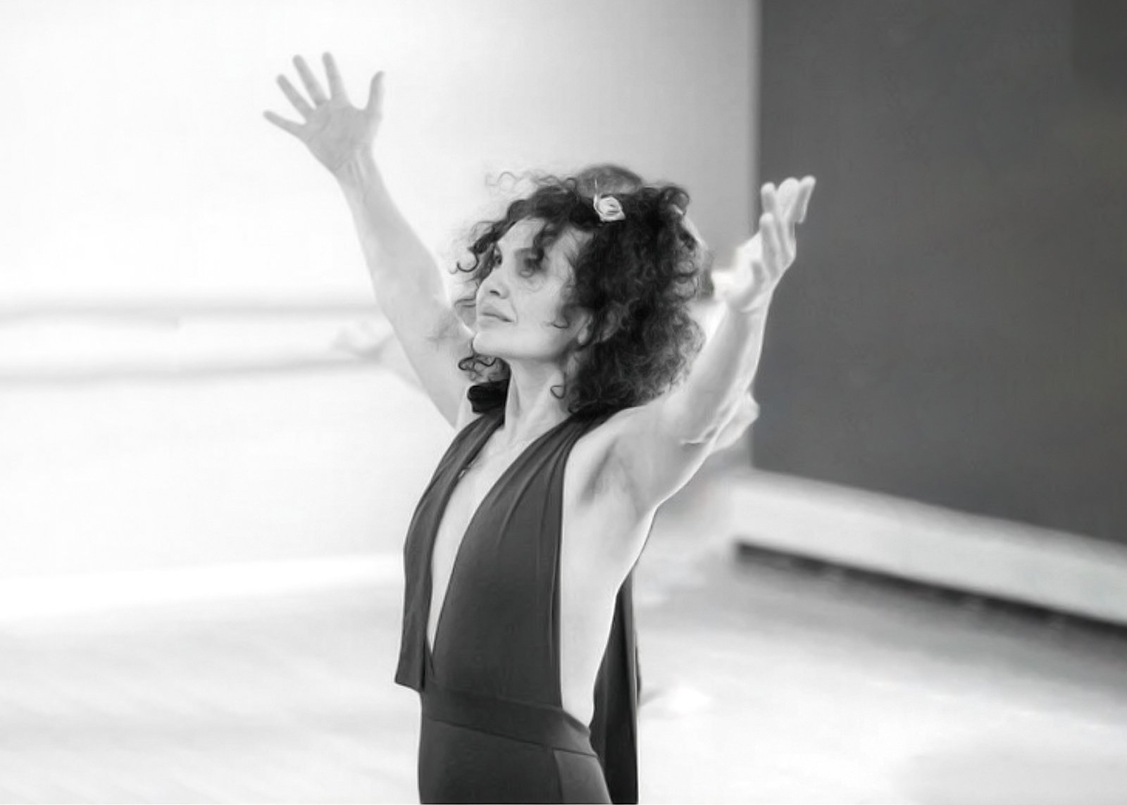 A woman with curly hair and a flower hair clip, wearing a sleeveless dress, raising her arms with her eyes closed in a dance studio.