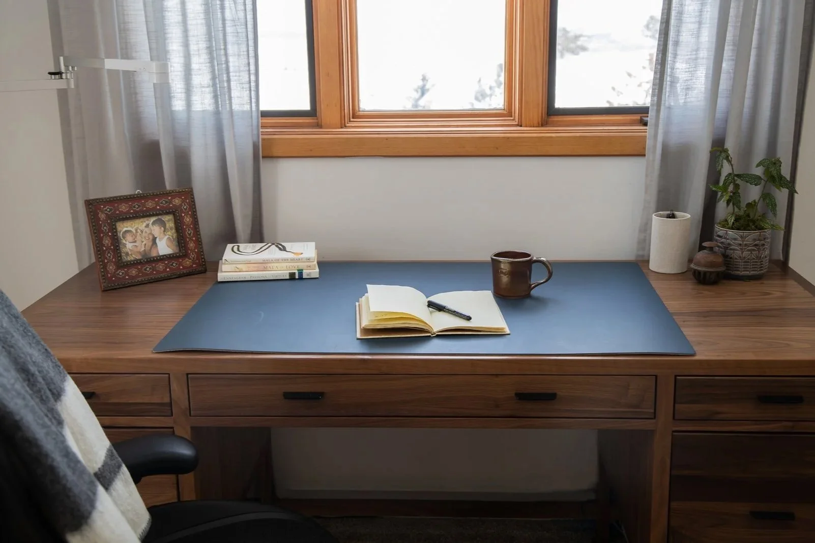 Wooden desk with open notebook, black pen, brown mug, and a blue desk pad, near a window with sheer curtains, with framed family photo, books, a potted plant, and decorative items on the side.