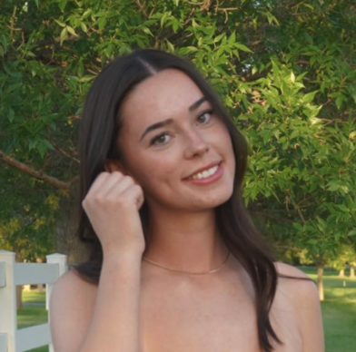 A young woman with long dark hair smiling outdoors in front of green trees, wearing a sleeveless top and a delicate necklace.