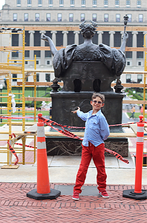 A young boy in red pants and a blue shirt is pointing at a large bronze statue of a historical figure, surrounded by orange traffic cones and red caution tape. The statue is behind a construction area with yellow safety barriers, in an urban setting with buildings in the background.