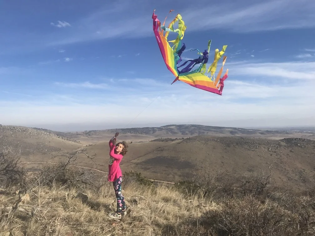 A girl in a pink sweater and floral pants flying a rainbow-colored kite in a dry, hilly landscape under a partly cloudy sky.