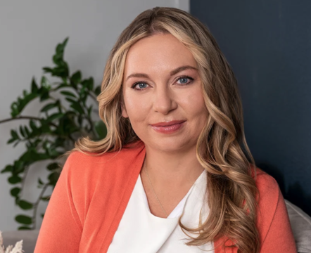Close-up of a man with dark curly hair, glasses, and a beard, wearing a dark green sweater, looking to the side with a slight smile, indoors with a light-colored wall and a plant in the background.