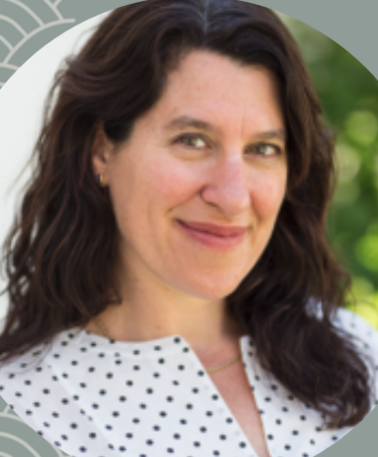 Close-up of a woman with wavy brown hair smiling outdoors, wearing a white top with black polka dots.