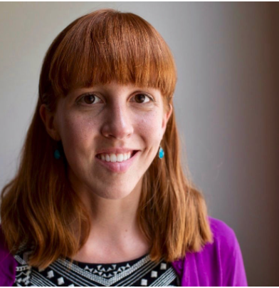 A woman with red hair, blue earrings, and a purple cardigan smiling at the camera.