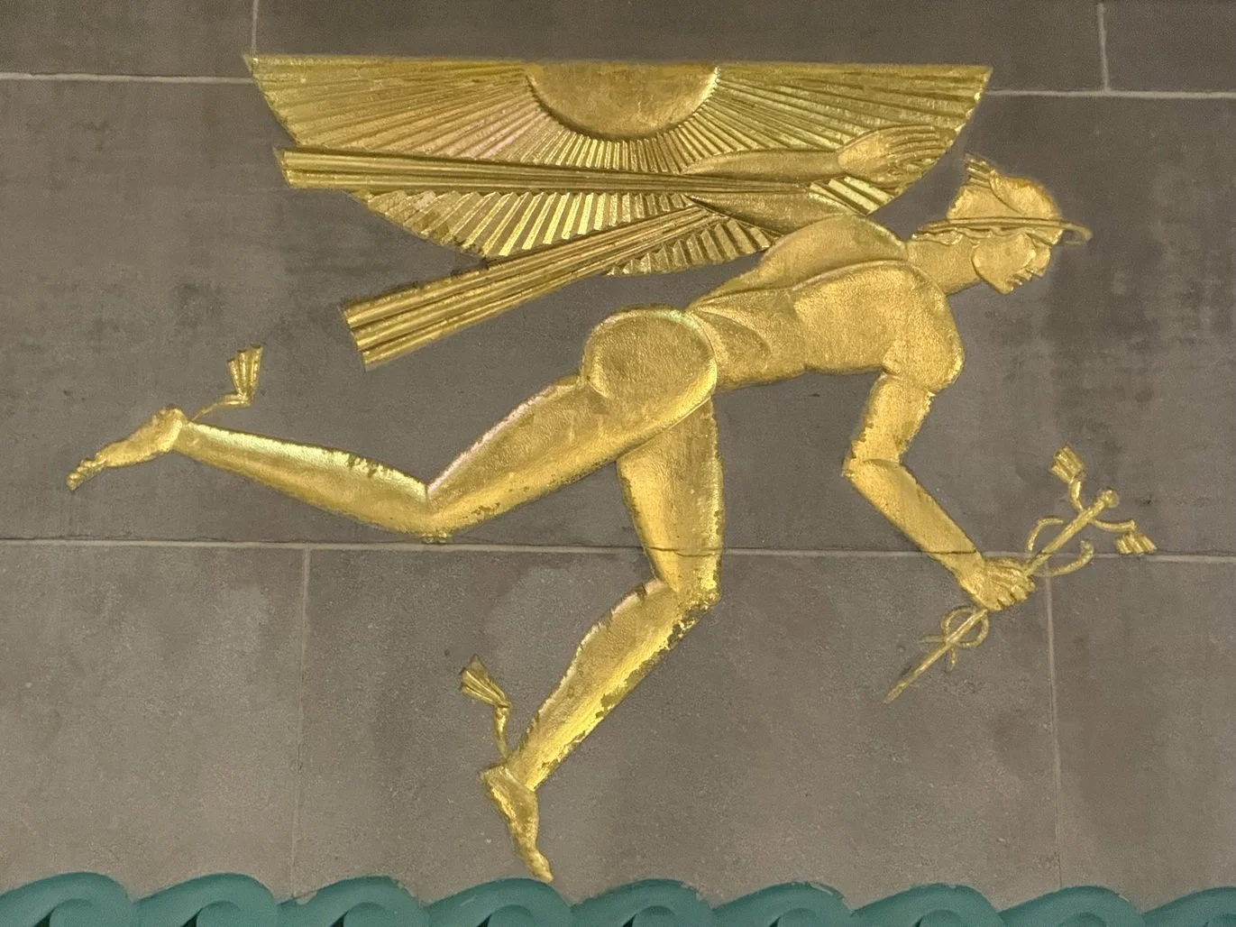 Gold-colored sculpture of a person running, carrying a large fan on their back and holding a staff in one hand.