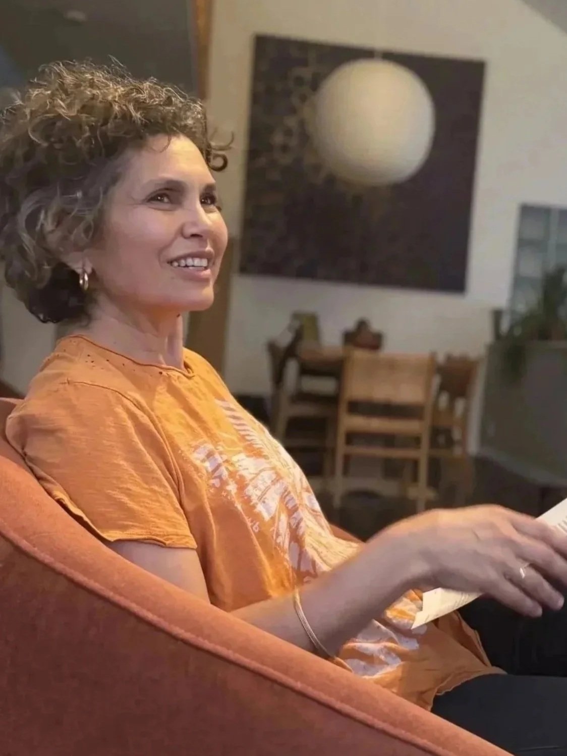 A woman with curly hair wearing a yellow-orange shirt, sitting on an orange chair, smiling, in a home interior with a dining table, chairs, and wall art.
