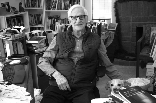 Matthew Fox  sitting at a cluttered desk in a room filled with books, with a dog sitting beside him.