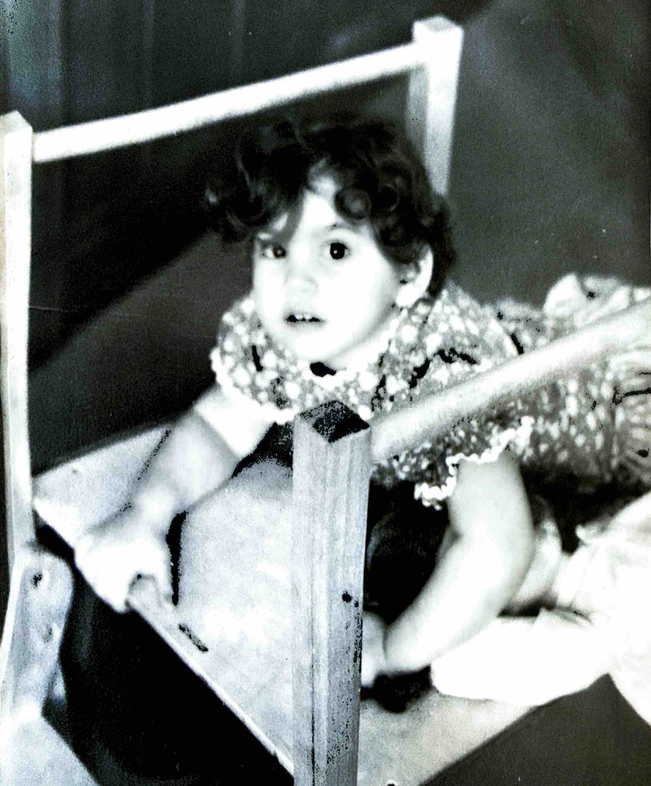 A young girl with curly hair in a patterned dress, looking up with an surprised or curious expression, inside a small wooden playpen or crib.