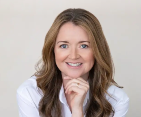 A woman with long wavy brown hair, blue eyes, wearing a white blouse, smiling, with her hand resting on her chin, against a plain light background.