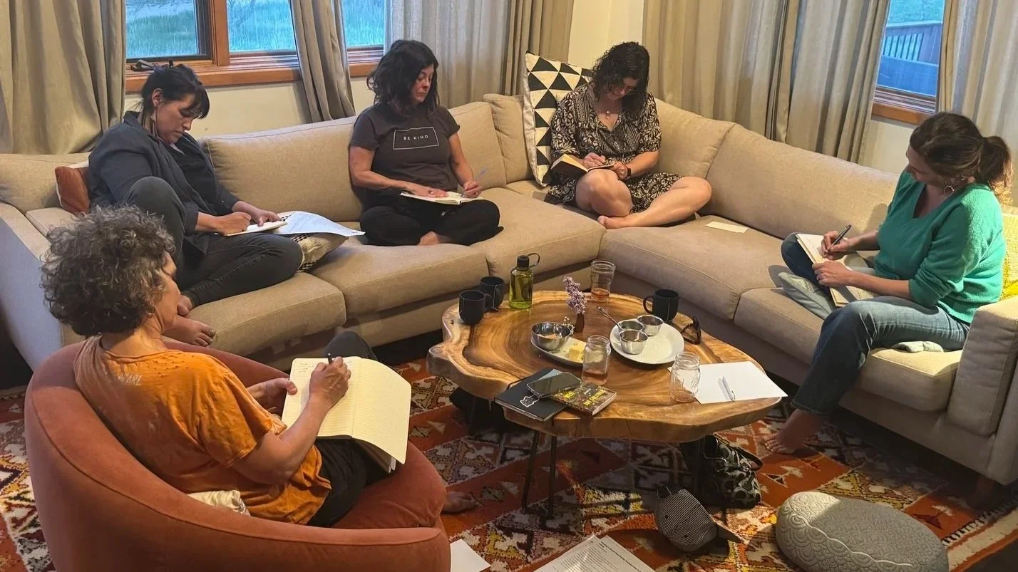Six women sitting around a coffee table in a living room in the midst of a writing circle in progress.