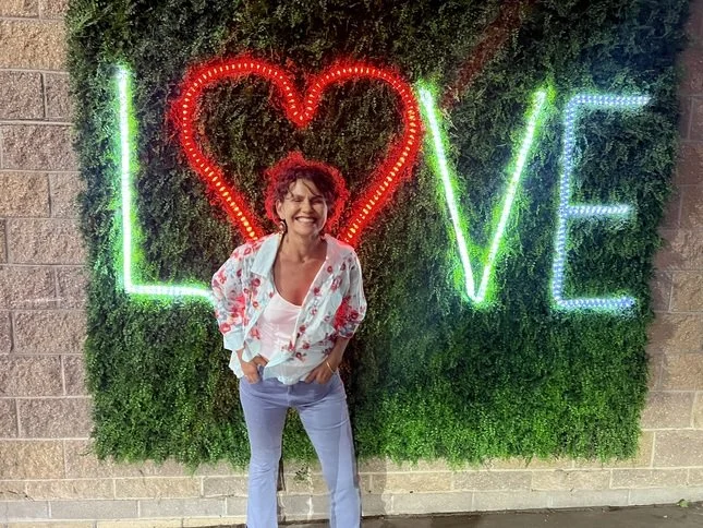 Woman standing in front of a decorative wall with neon lights spelling 'LOVE' and a heart, smiling and posing.