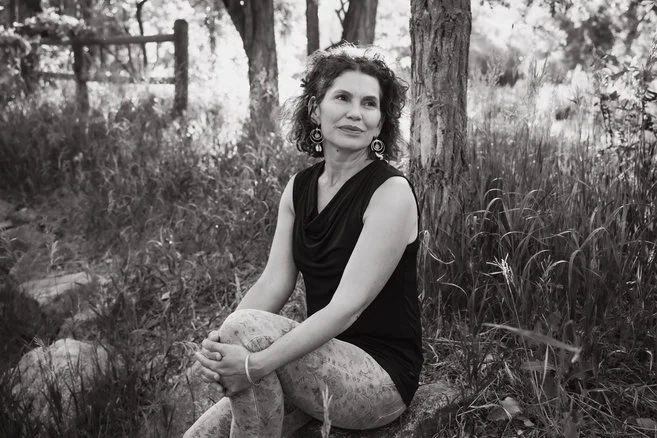 A woman with curly hair sitting on the ground outdoors near a tree.