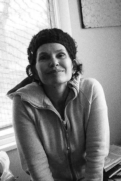 Black and white photo of a woman wearing a hoodie and a knit cap, sitting indoors near a window.