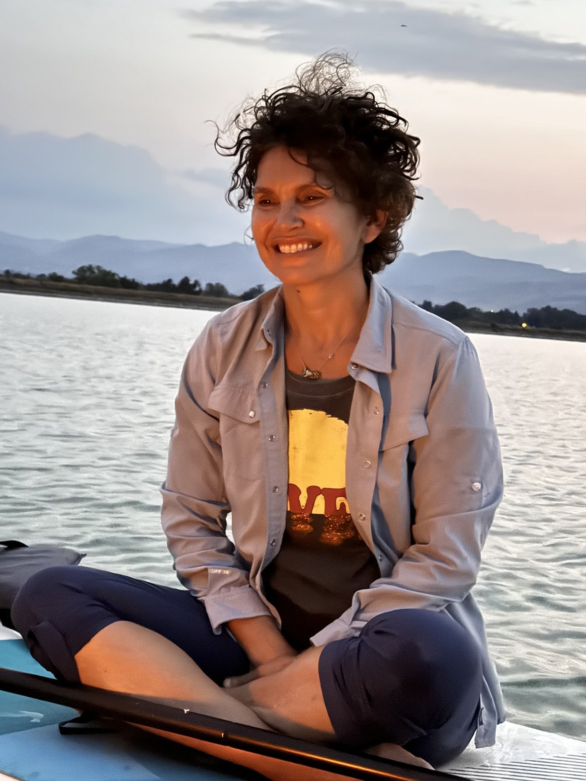 A woman with curly hair sitting cross-legged on a paddleboard on a body of water during sunset, smiling and wearing a gray shirt over a graphic t-shirt.