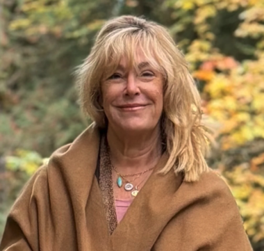 A man with curly dark hair, a beard, and glasses, wearing a dark green sweater, looking to the side with a neutral expression inside a room with a white wall and a plant.