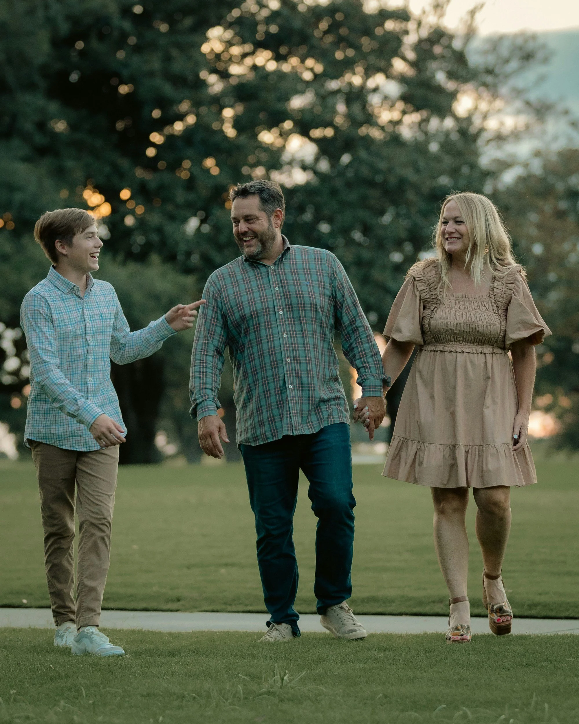 A family of three walking outdoors on a grassy area during sunset, holding hands and smiling, with trees and a sunset in the background.