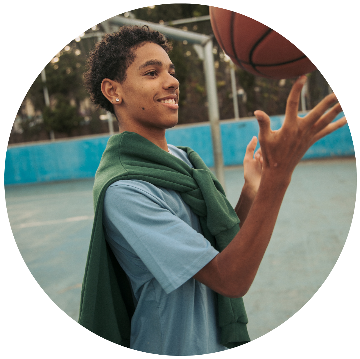A young person with short curly hair spinning a basketball on their finger on an outdoor court, smiling and looking to the side.