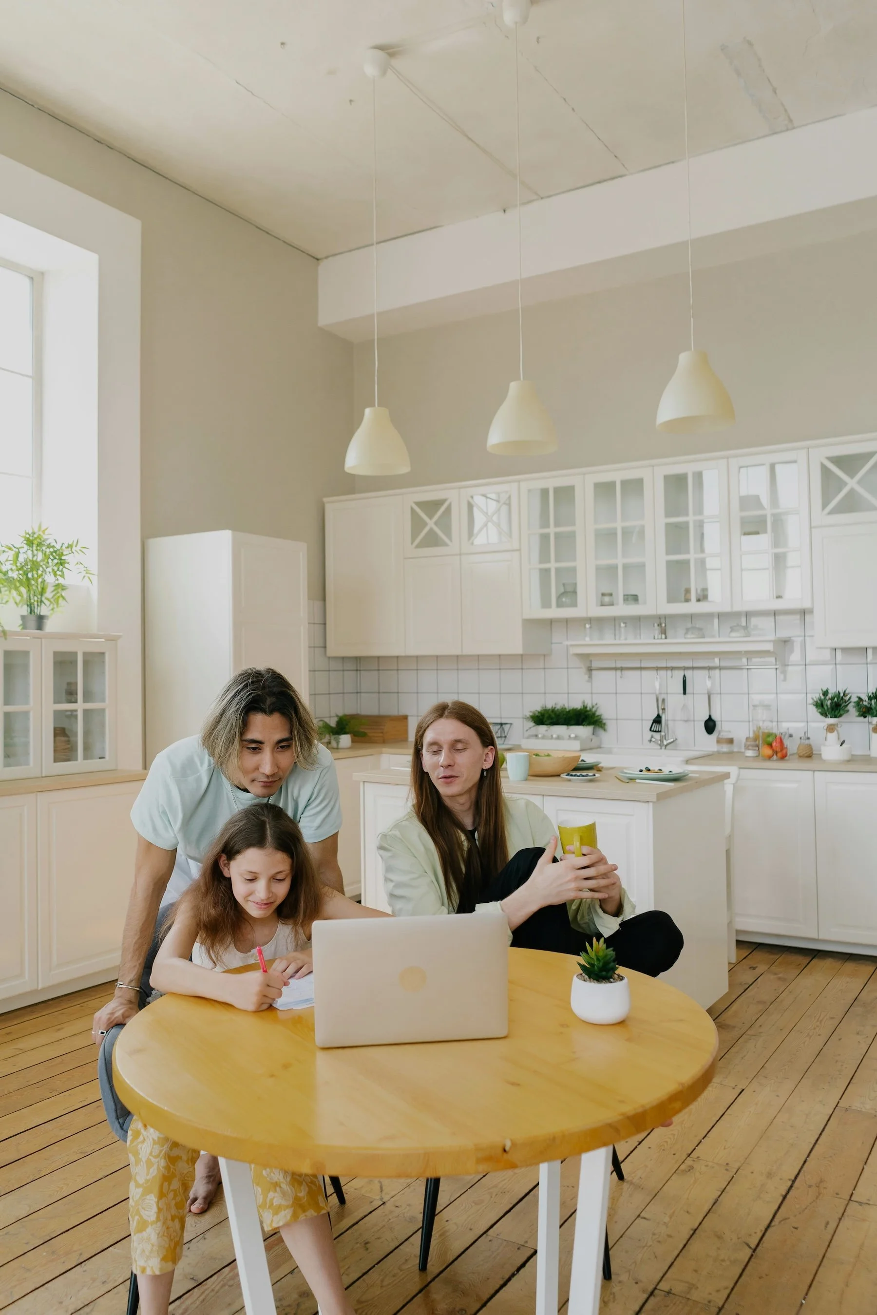 A family of three sitting at a round wooden table in a white kitchen, with the father and daughter focused on a laptop and the mother holding a mug, all engaged in a shared activity.