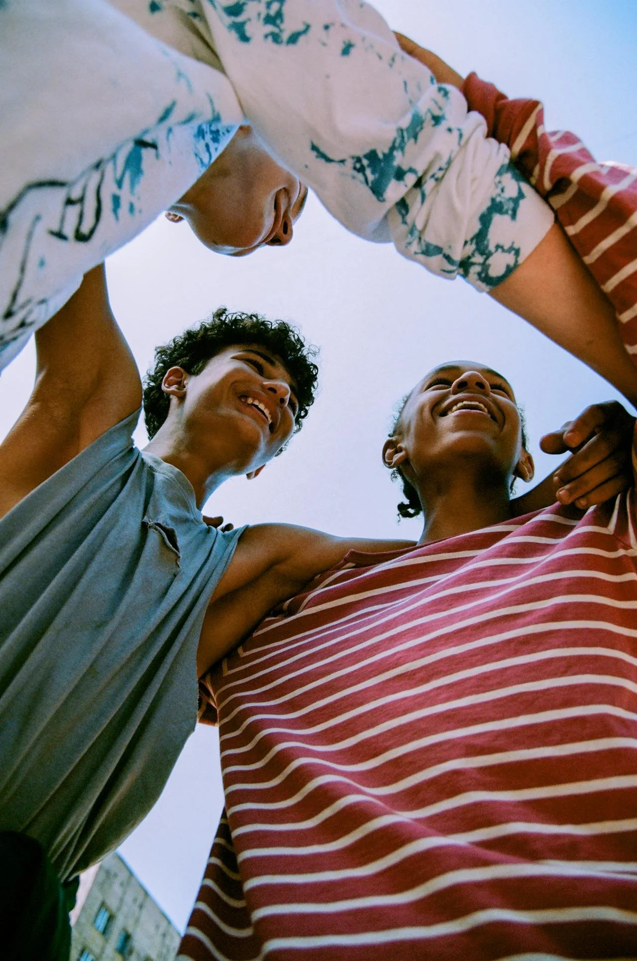 Three young people forming a circle with their arms raised, smiling and looking at each other, taken from below against a clear sky.