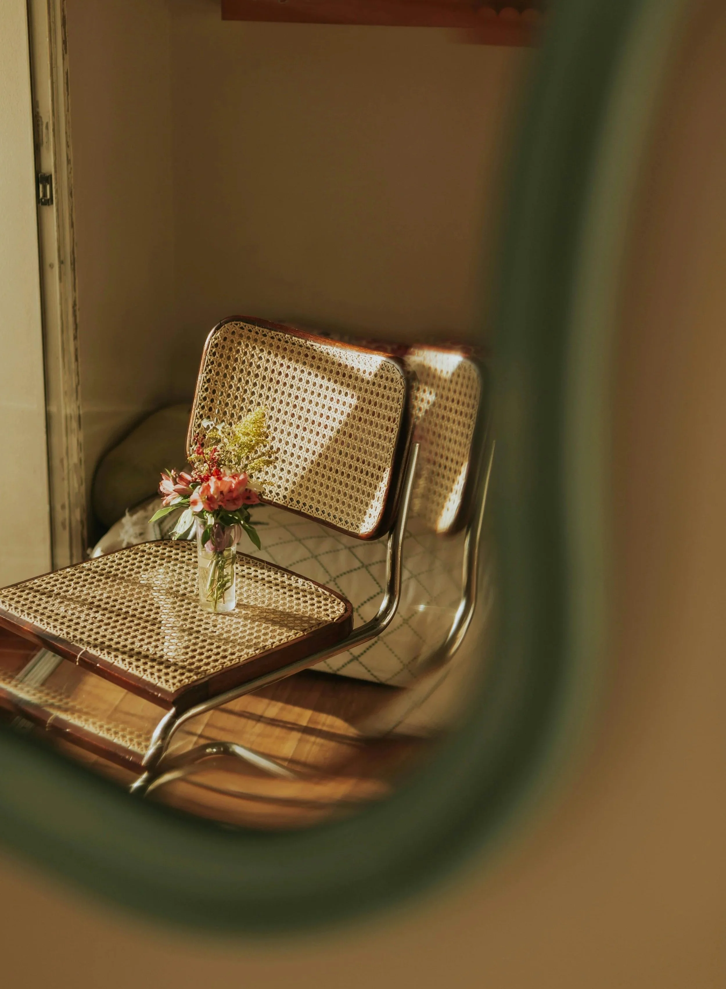 brown chair in a sunlit room with a hardwood floor and white walls. There is a small vase on the chair with pink and yellow flowers.