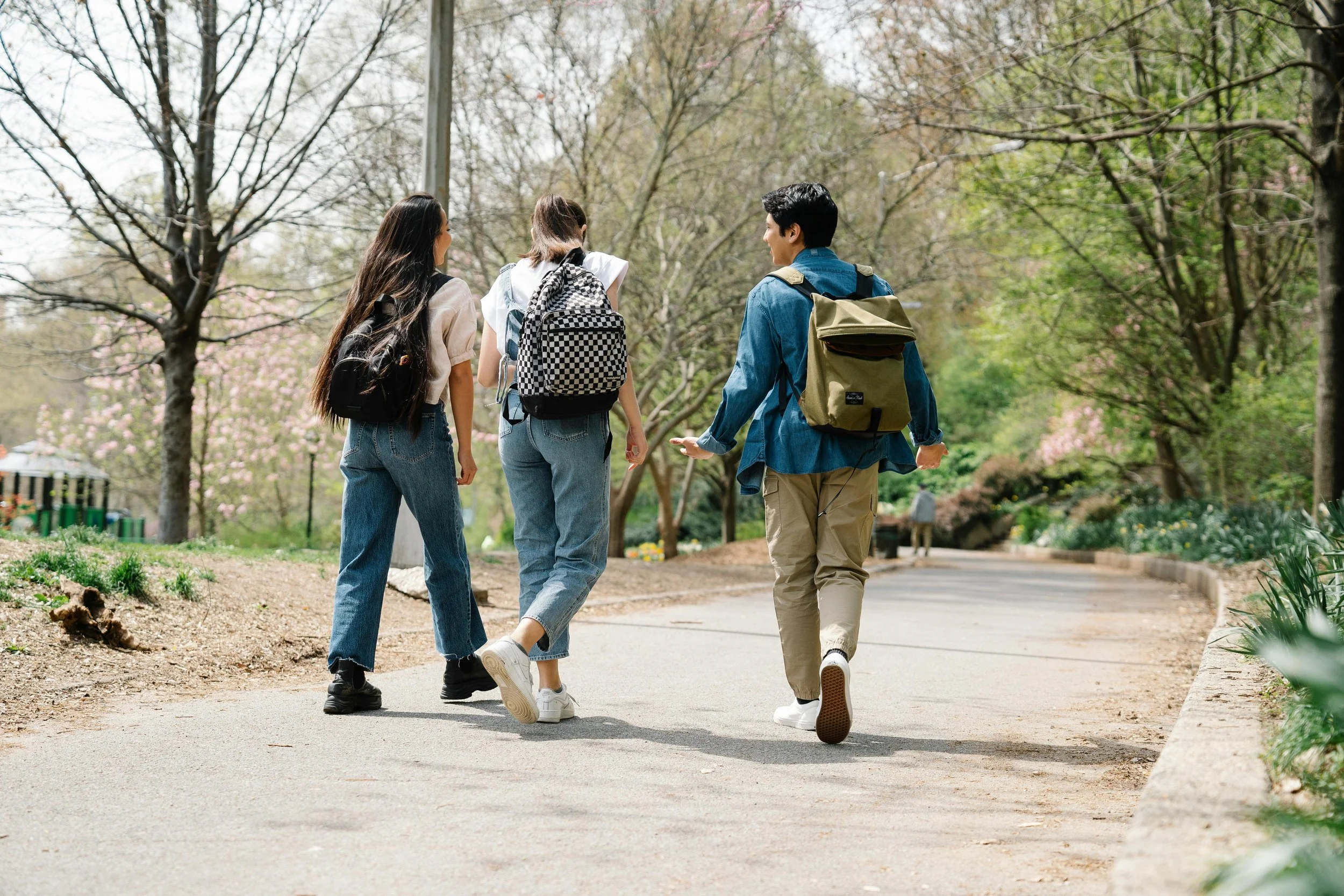 Four young people walking and talking along a park pathway surrounded by trees and blooming flowers, carrying backpacks.