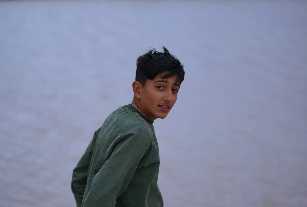 A teenage boy with short dark hair and a slight smile, wearing a green jacket, standing against a pale, cloudy sky background.