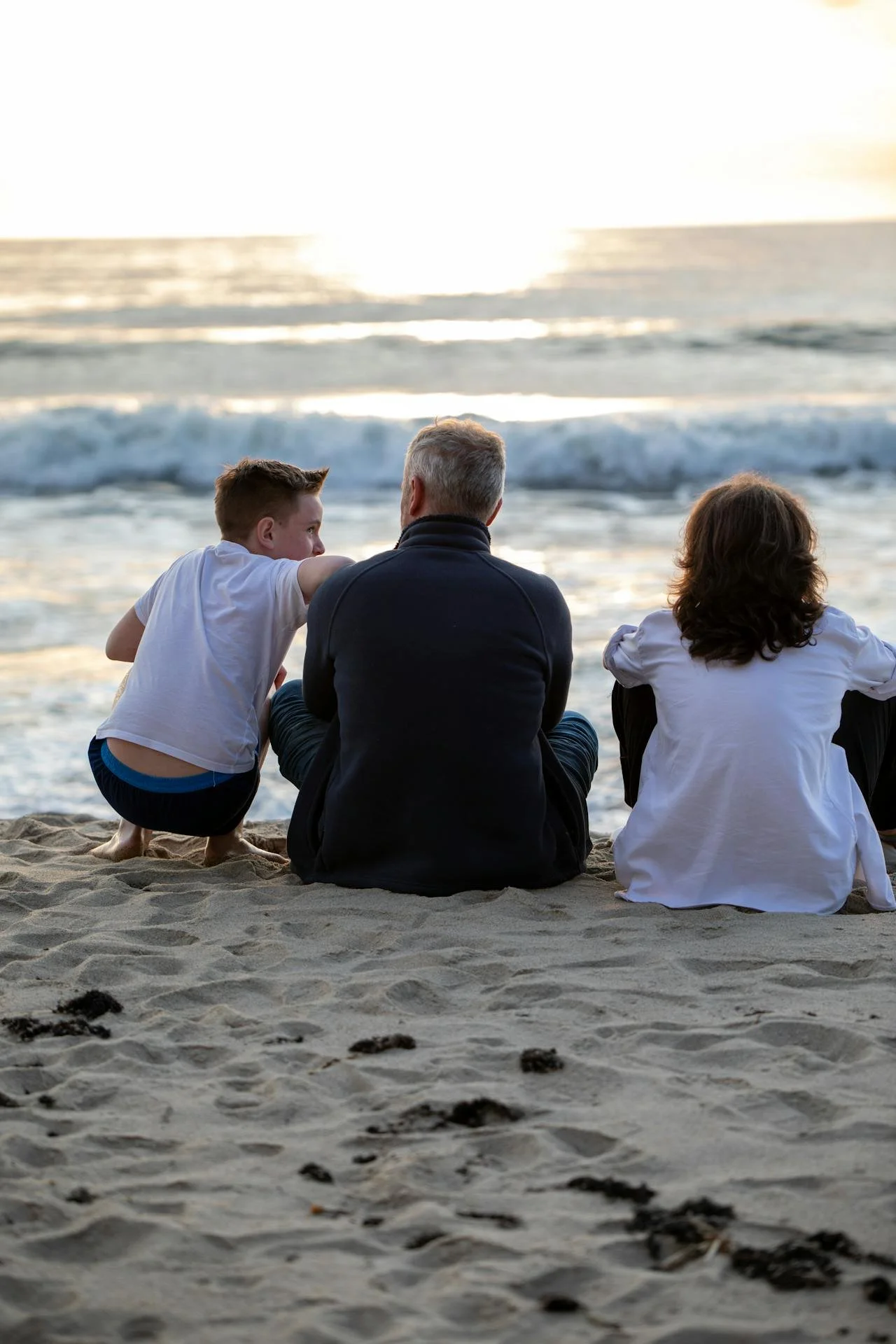 A family of three sitting on the sandy beach and looking at the ocean waves during sunset.