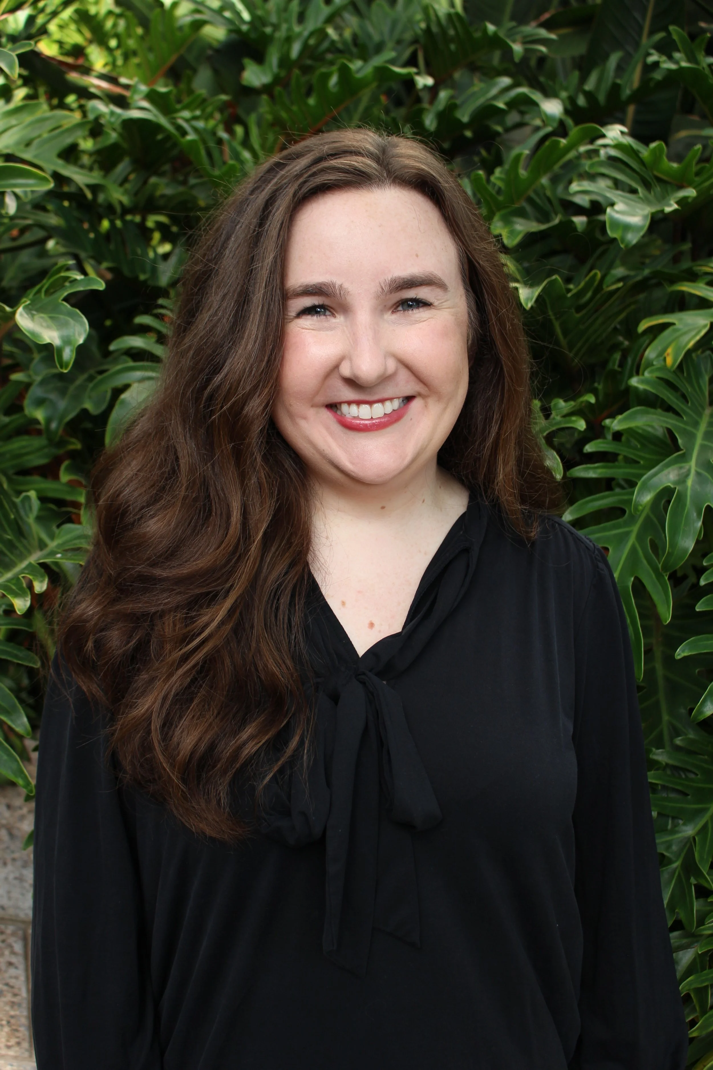 portrait of Dinah Farrell against a leafy green background. She is wearing a dark v-neck shirt and a necklace with a pendant. She has light skin, a big smile and long wavy brown hair.