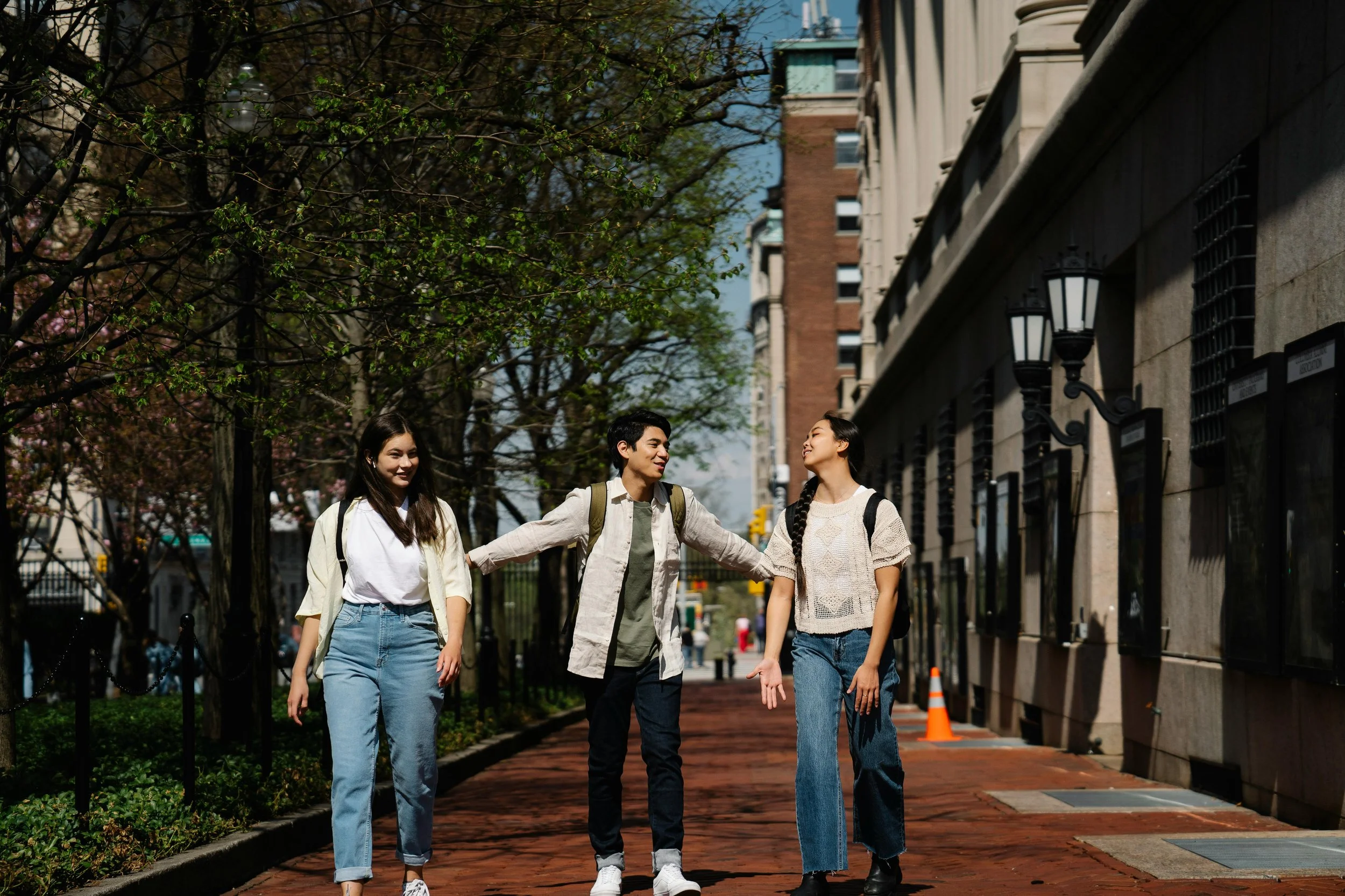 a young man and a young woman walking, talking  anon a city sidewalk on a sunny day, with trees and urban buildings in the background. They are smiling and both wearing backpacks.