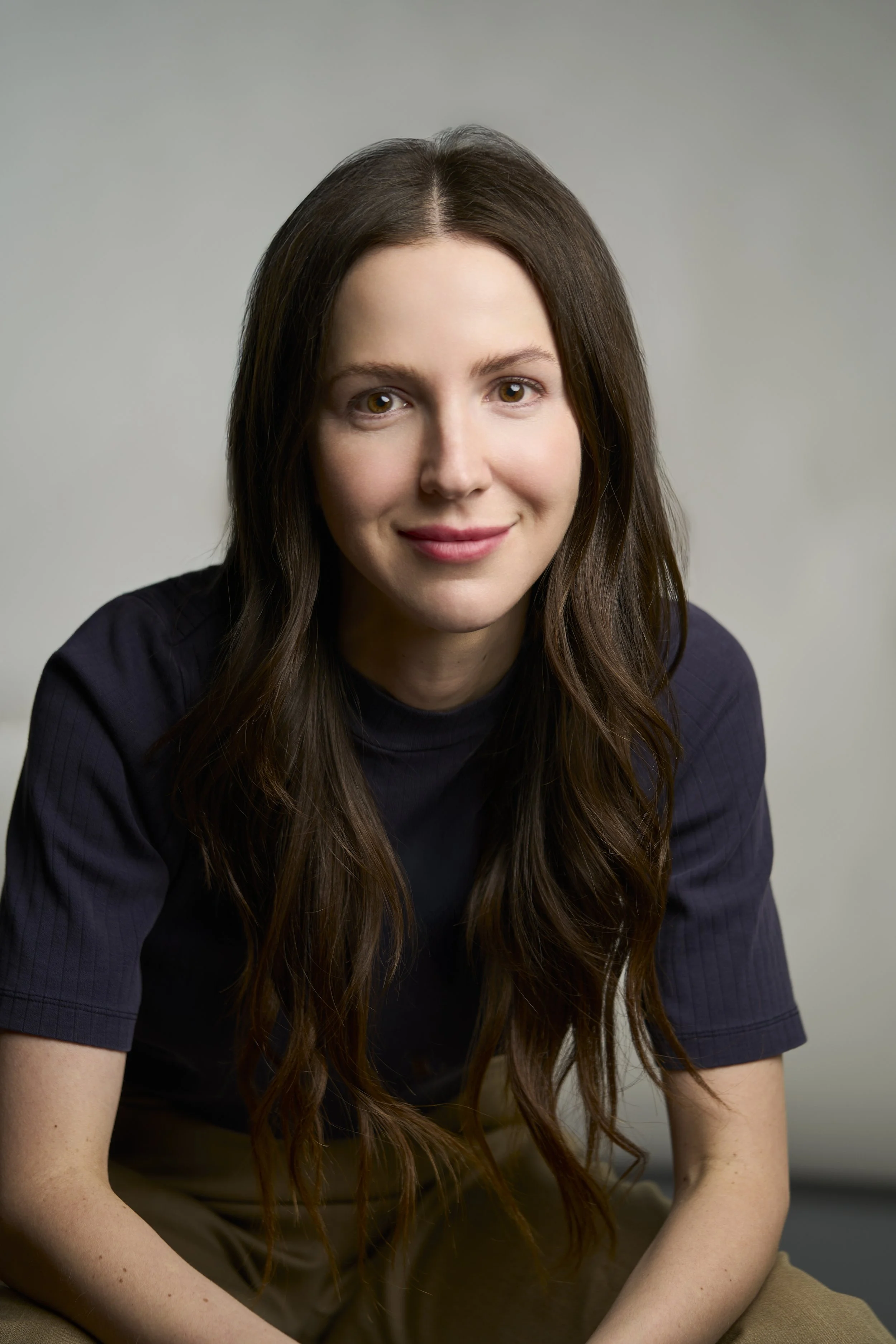 Portrait of a Dr. Sam Harris. She has light skin and long wavy dark brown hair, wearing a dark short-sleeved top and beige pants, smiling softly at the camera against a plain gray background.