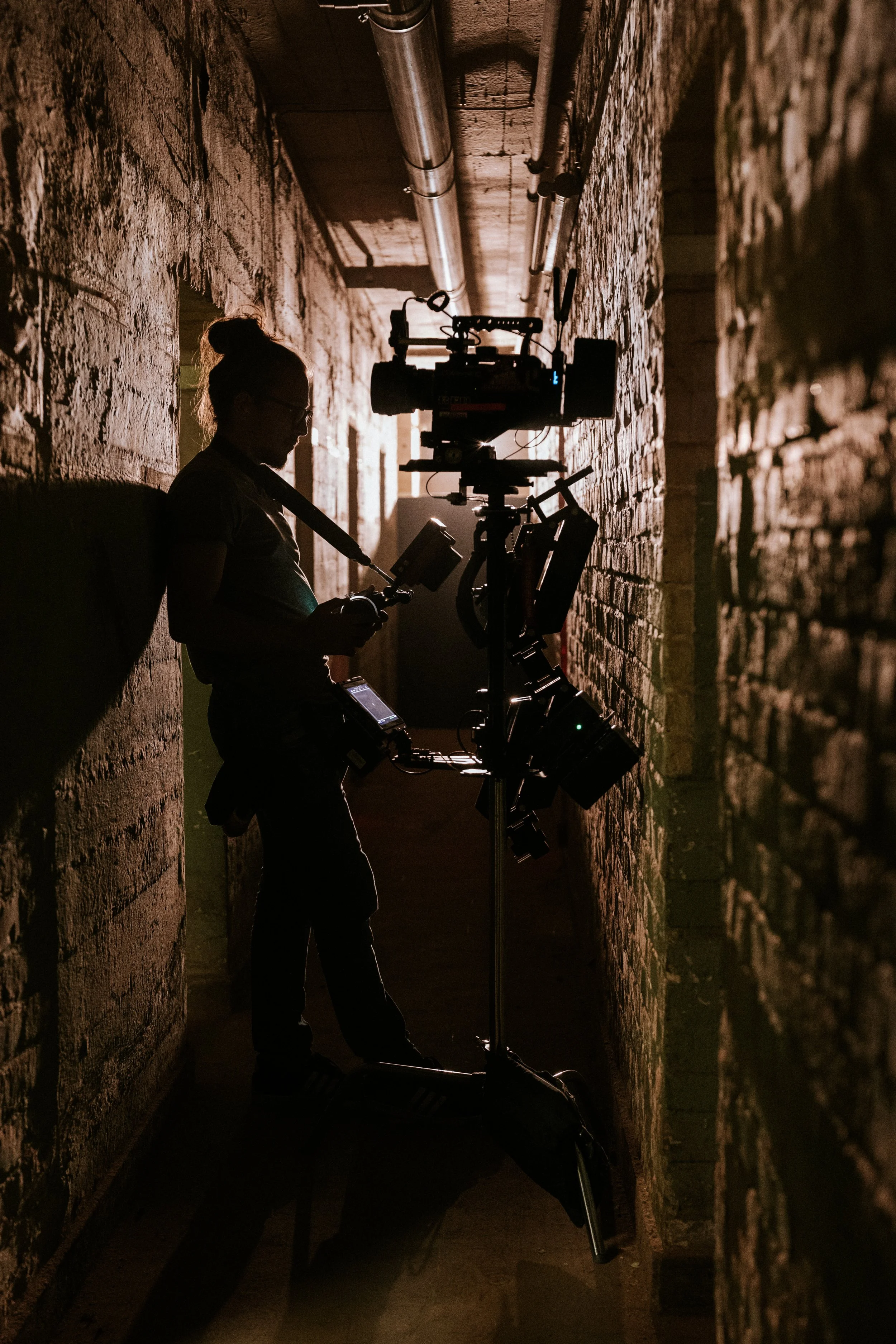 Eine Person, wahrscheinlich ein Filmemacher oder Fotograf, steht mit Kamera und anderem Equipment in einem dunklen, engen Flur mit steinernen Wänden, beleuchtet nur durch eine Lichtquelle im Hintergrund.