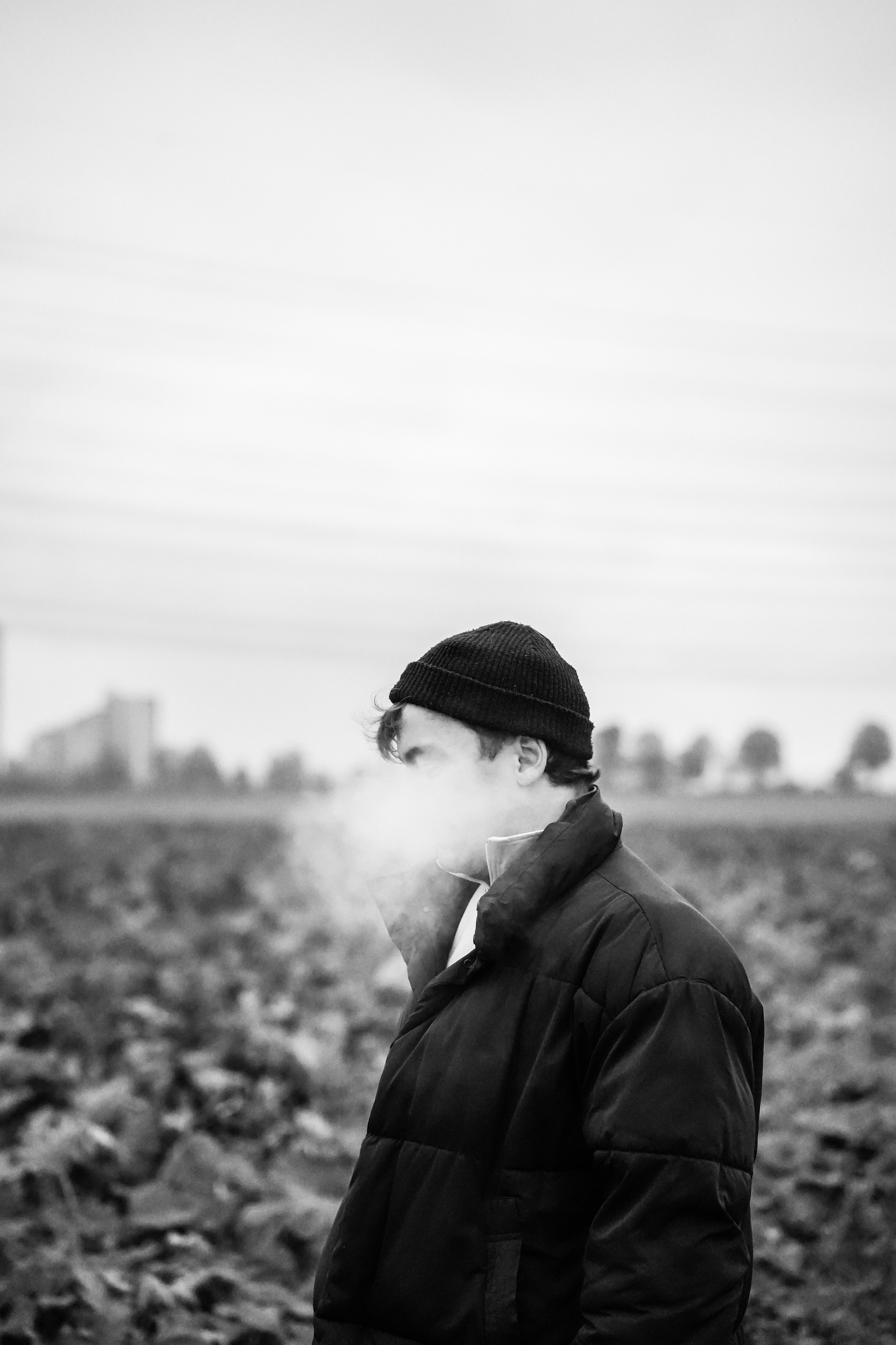 Junge in dunklem Jacke und schwarzen Mütze raucht in landwirtschaftlicher Umgebung, Zeuge von Nebel oder Rauch. Schwarz-Weiß-Foto, verschwommene Gebäude im Hintergrund.
