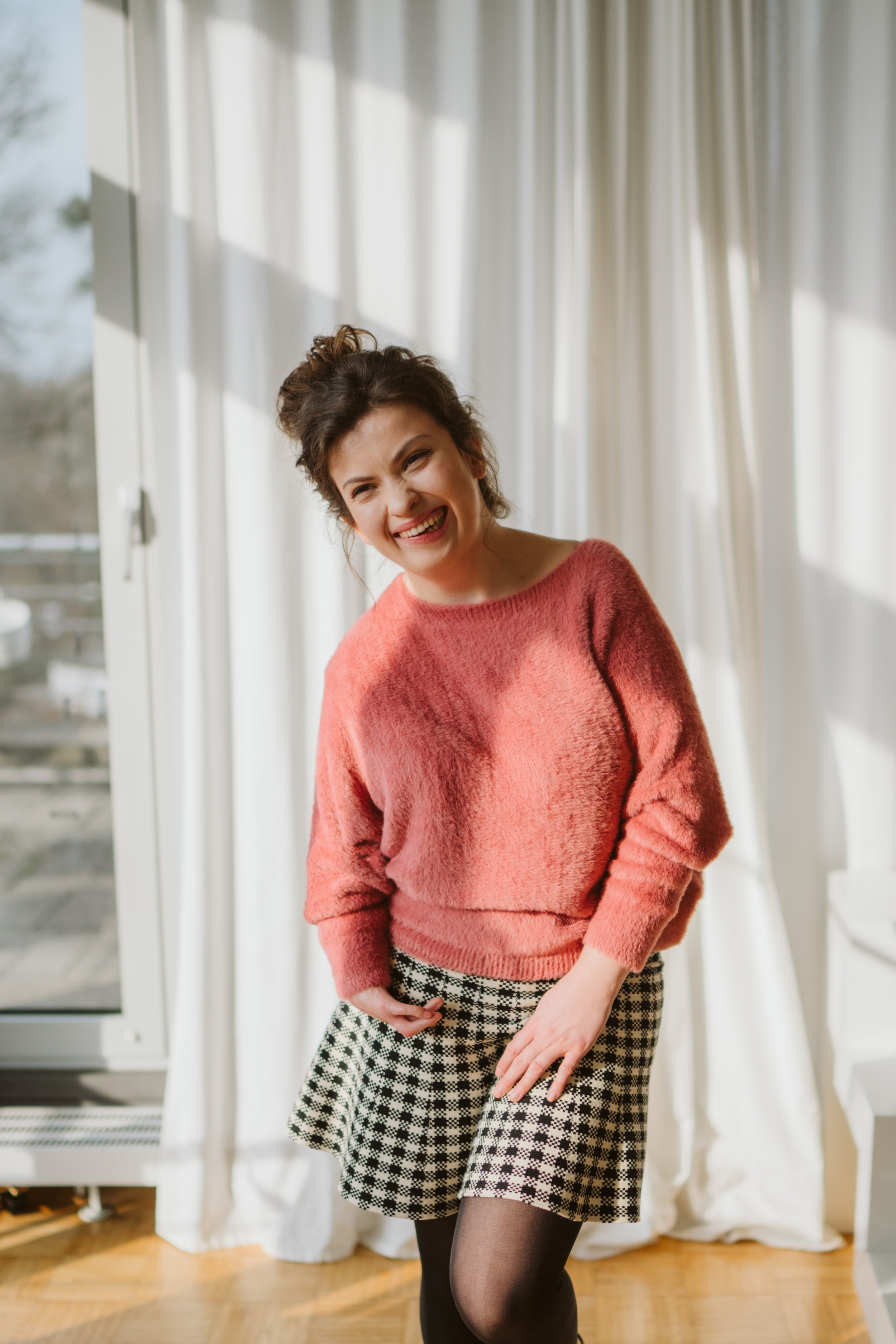 Junge Frau lachend in einem hellen Zimmer, trägt roten Pullover und schwarz-weiß karierte Shorts, im Hintergrund Vorhänge und ein Fenster.