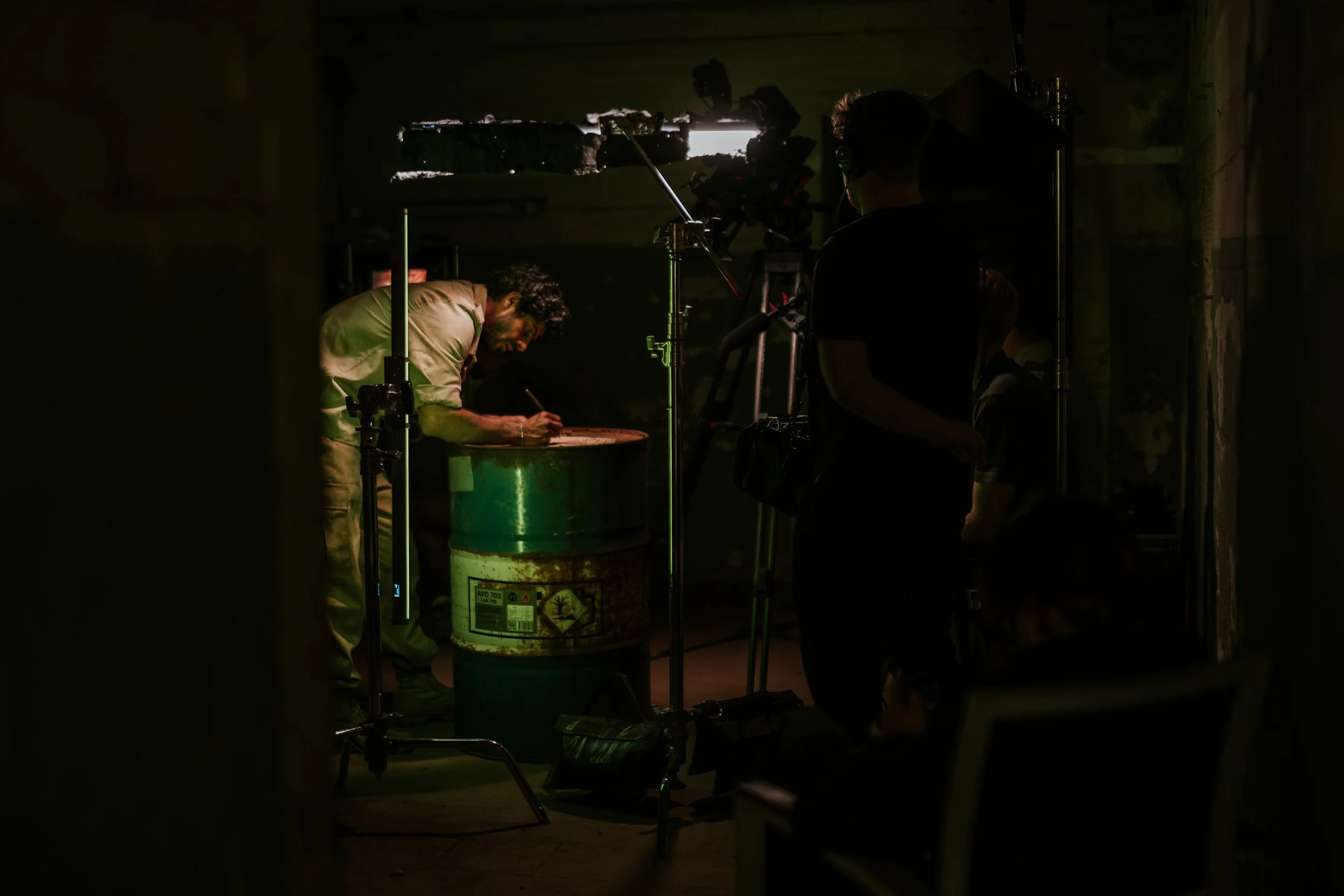 Dreharbeiten mit einer Person, die an einem grünen Metallfass schreibt, in einem dunklen Raum mit Filmtechnik und Licht. Zwei andere Personen sind silhouettenhaft und beobachten.