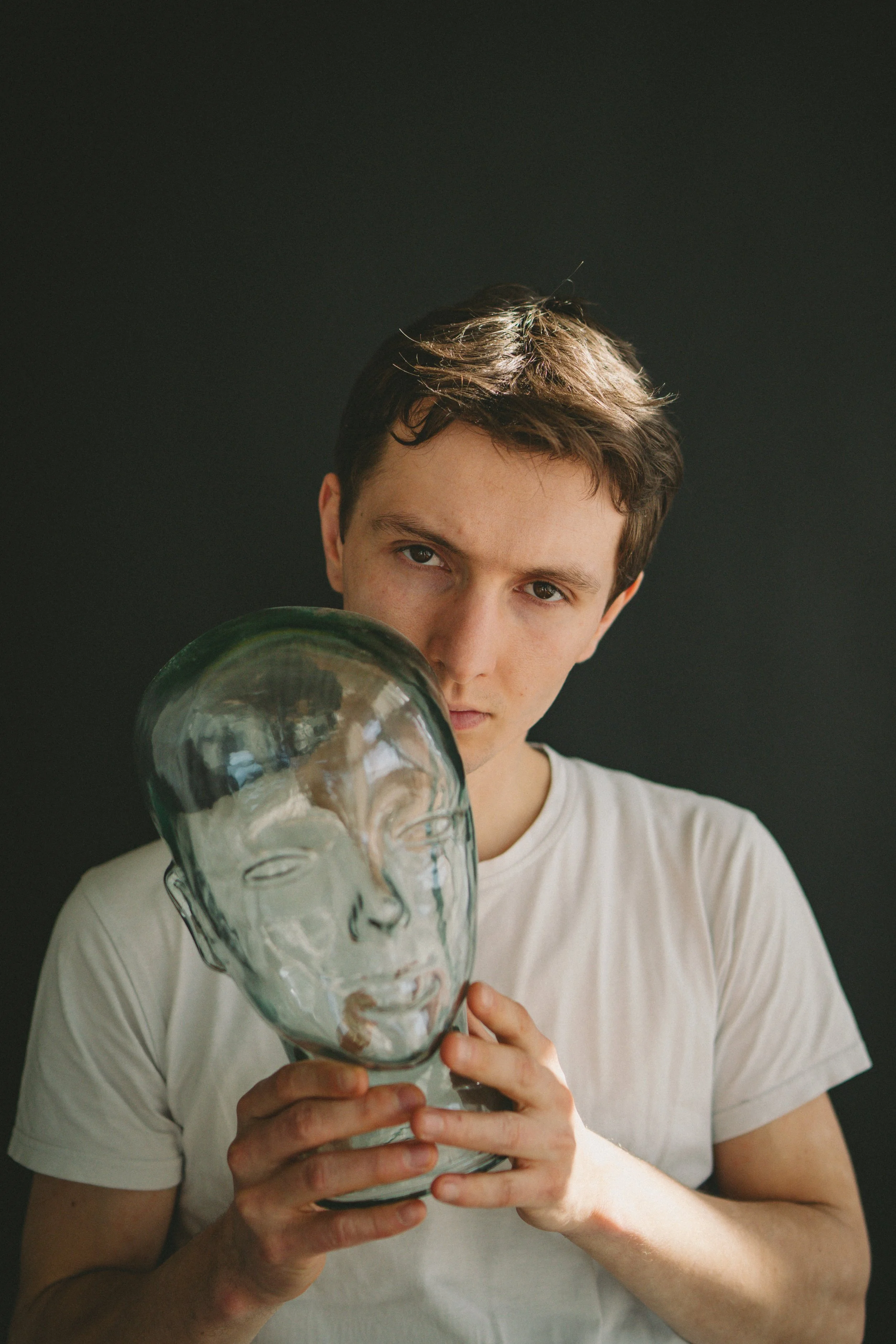 Junge hält eine transparente Glasplastikmaske eines menschlichen Gesichts vor die Brust, gegen einen dunklen Hintergrund.