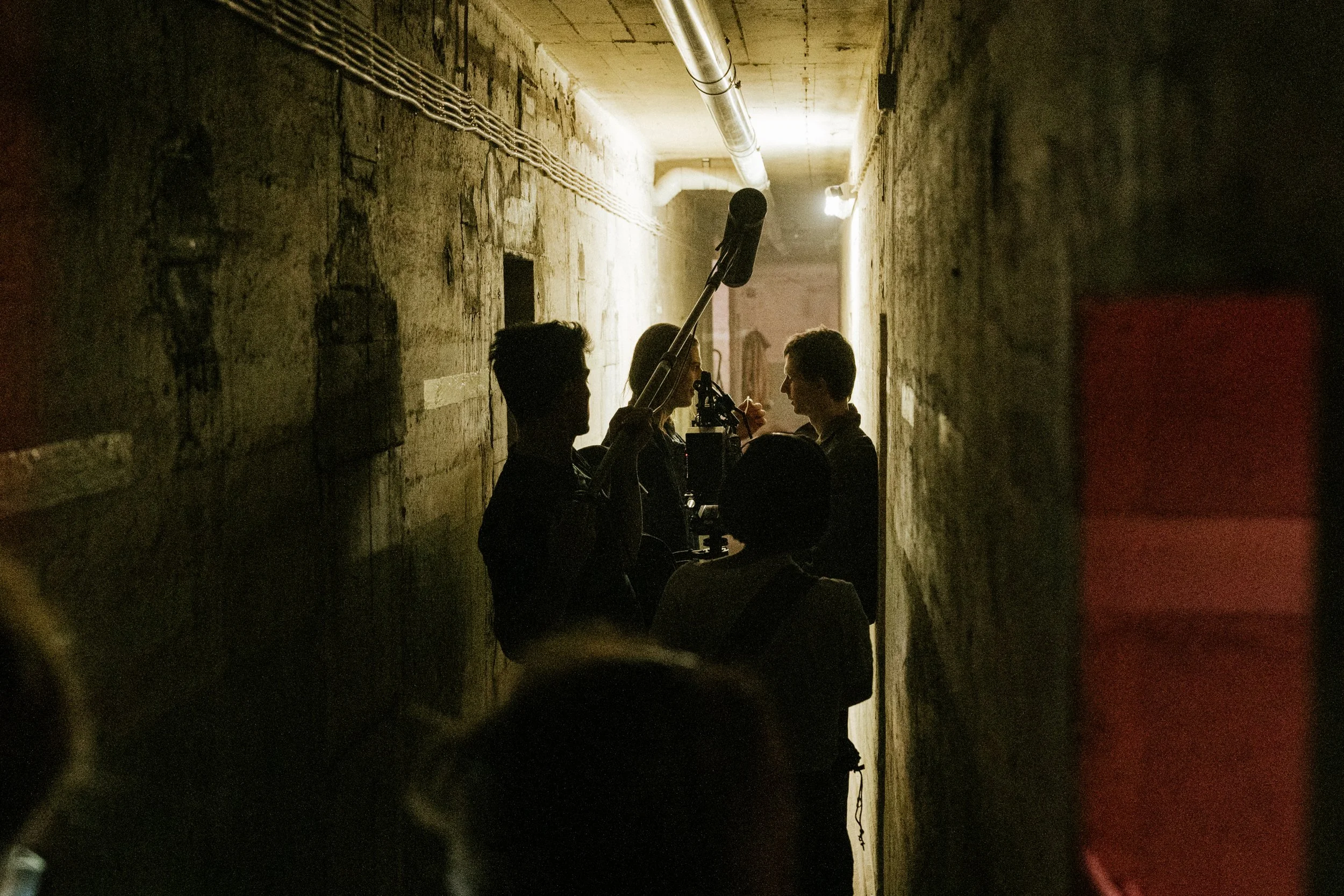 Drei Menschen in einem engen, dunklen Flur mit rauen, alten Wänden. Zwei von ihnen sprechen miteinander, während die dritte Person eine Tonaufnahme mit einem Mikrofon macht.